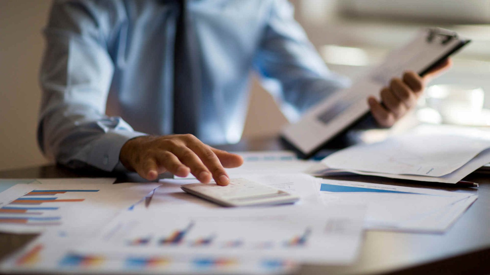 An accountant, with papers spread across his desk, types into a calculator and holds a clipboard as he calculates something 