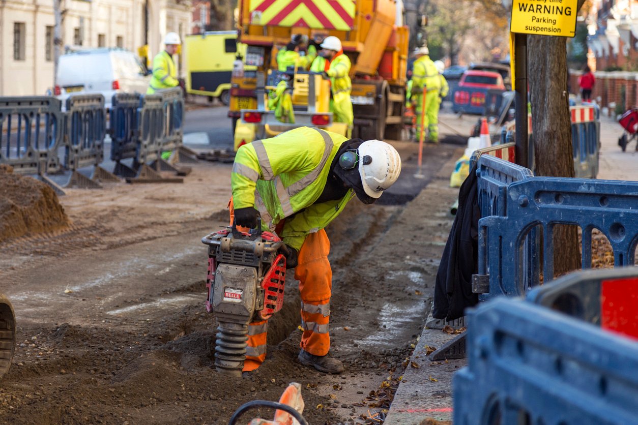 Construction worker using equipment at a roadwork site, with colleagues in the background, all wearing high-visibility gear—tracked using Causeway SkillGuard for compliance and safety