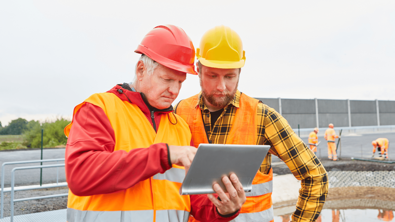 Construction worker in an orange safety vest holding a coffee cup and tablet on a job site with tools and blueprints nearby - Causeway Skillguard