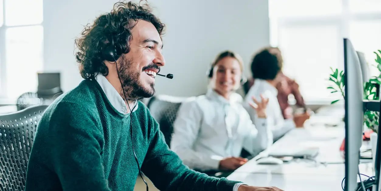 A male construction software support worker sits at his desk and laughs as he talks into a headset. A female colleague does the same behind him