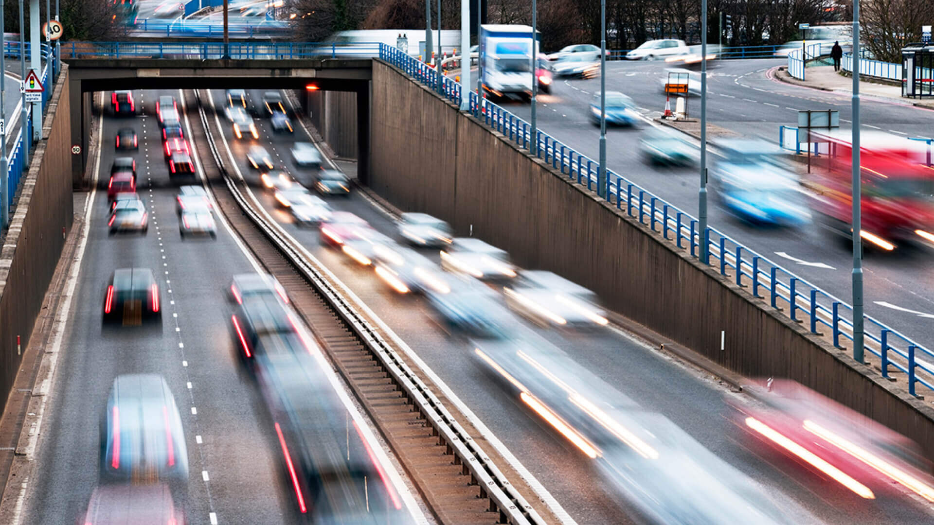 Blurred motion of vehicles on a busy multi-lane urban motorway near Birmingham with an underpass, surrounded by barriers, streetlights, and adjoining roads in the background. Birmingham is the location of the ITS UK and Causeway D-TRO event.