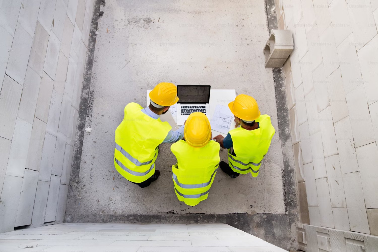 Top view of three construction workers in yellow safety vests and hard hats reviewing plans on a laptop at a building site - Causeway Skillguard