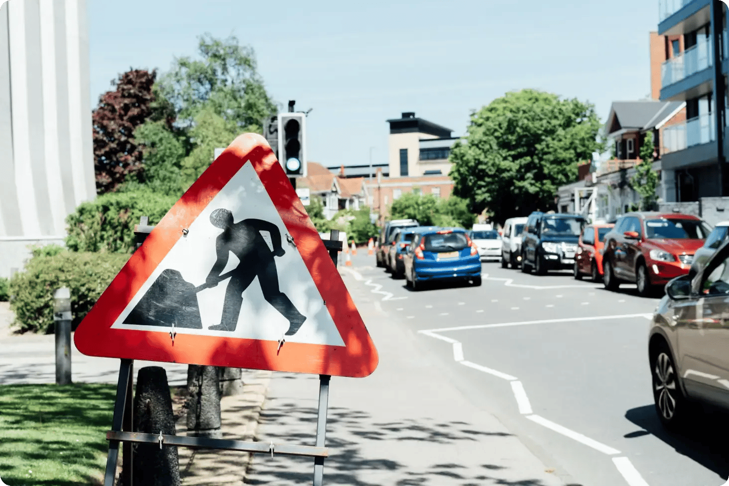 Roadworks sign on the the left hand side of UK road. Cars queuing at traffic lights.